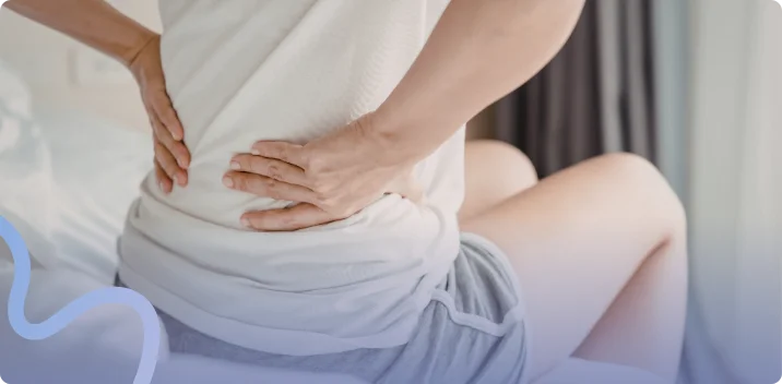 Woman sitting on the edge of a bed pressing both hands into her aching lower back