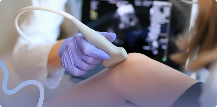 Clinician in gloves guiding an ultrasound therapy probe along a patient's knee with the monitor in the background
