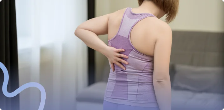 Woman in a purple athletic top reaching behind her to hold her aching lower back at home